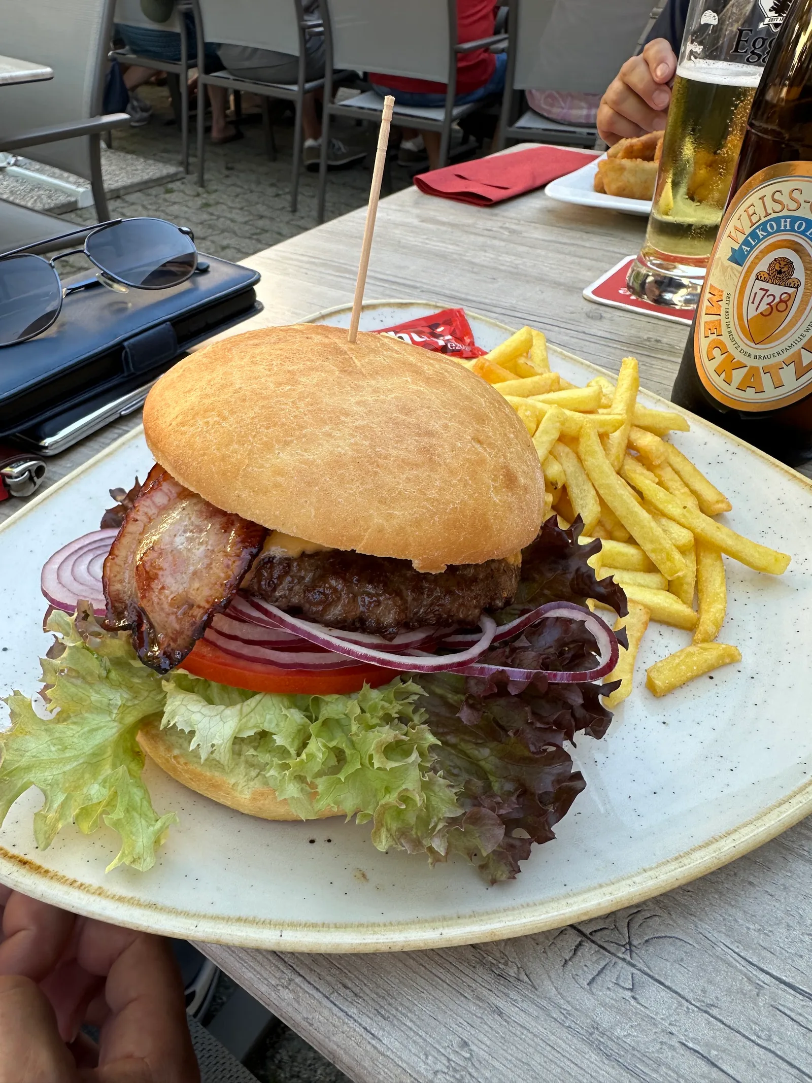 Burger mit Speck, Salat und Pommes frites auf einem Tisch im Gastgarten, daneben ein Glas Bier