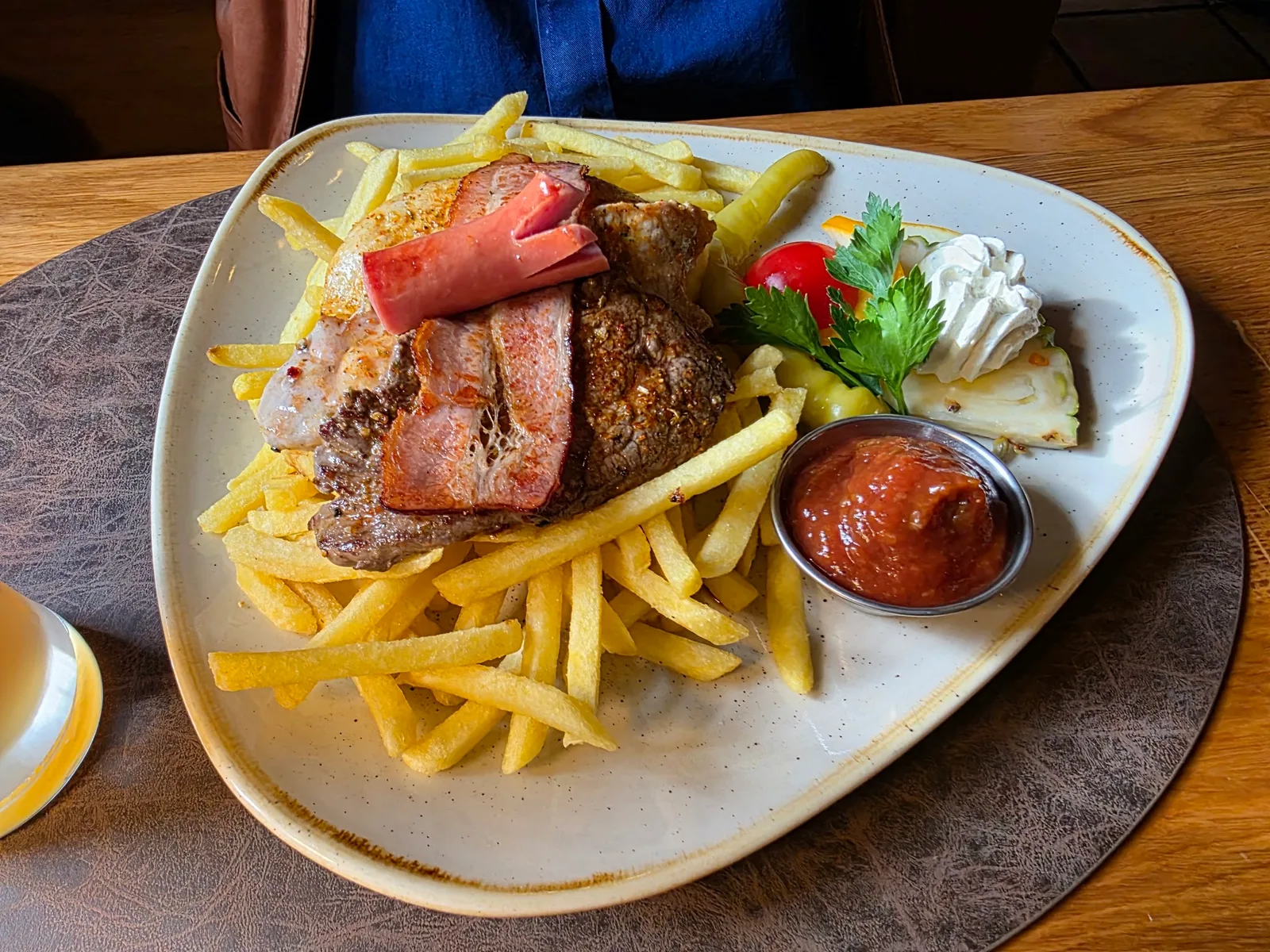 Rostbrätl mit gebratenem Speck, Pommes frites und einem Schälchen Sauce auf hellem Teller